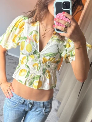 Blommig croppad blus med citroner - Söt croppad blus med blommigt och citronmönster i vitt, gult och grönt. Blusen har puffiga korta ärmar, v-ringning och smock i midjan för en snygg passform. Perfekt för sommarens alla tillfällen!