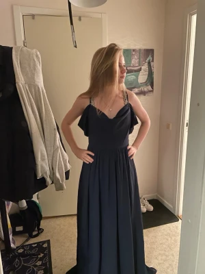 Marinblå bal  långklänning med volang - Säljer en elegant marinblå bal  långklänning med volangdetaljer vid axelbanden och v-ringad fram och bak. Klänningen har smala axelband och ett flödande fall som ger en snygg siluett. Perfekt för dig som vill ha en stilren look.