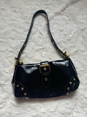 Svart axelremsväska med nitar - Snygg svart handväska i glansigt material med axelrem och guldfärgade nitar. Väskan har ett spänne framtill och detaljer i guld på både rem och kanter. Perfekt för dig som gillar en edgy och trendig look.