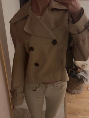 Trenchcoat hm - Trendig kort trenchcoat i beige med breda slag och dubbelknäppning framtill. Jackan har stora svarta knappar och justerbara ärmslut. Perfekt för dig som gillar en stilren och modern look. Materialet ser ut att vara bomullsblandning.
