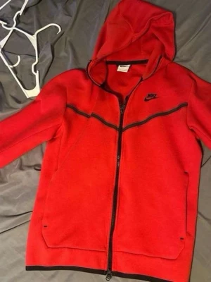 Röd Nike hoodie med dragkedja - Snygg röd hoodie från Nike med hel dragkedja framtill och svarta detaljer. Hoodien har huva, två fickor och en extra ficka med dragkedja på ärmen. Nike-logga i svart på bröstet och coola svarta linjer över bröstet. Perfekt för en sportig och avslappnad stil.