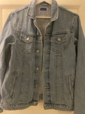 Ljusblå jeansjacka från Jack & Jones - Snygg ljusblå jeansjacka från Jack & Jones med klassisk krage, två bröstfickor med lock och knappar samt två sidofickor. Jackan har silverfärgade knappar framtill och detaljerade sömmar för en tidlös look. Perfekt att slänga över en hoodie eller t-shirt.