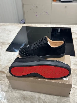 Svarta Louboutin sneakers med nitar - Snygga svarta sneakers från Christian Louboutin i mocka med ikoniska röda sulor och svarta nitar på tån. Skorna har lågt skaft, svarta skosnören och en lyxig känsla. Perfekta för dig som vill sticka ut med exklusiva detaljer och streetstyle-vibe.