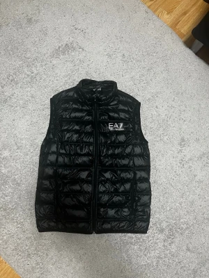 Svart dunväst från EA7 Emporio Armani - Säljer en svart, quiltad dunväst från EA7 Emporio Armani. Västen har hög krage, dragkedja framtill och en stilren logga på bröstet. Perfekt att slänga över en hoodie eller tröja för en sportig och clean look.