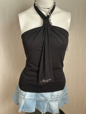 Svart halterneck topp från Morgan - Snygg svart halterneck topp från Morgan med dekorativ knytning i nacken och ett långt tygstycke framtill. Toppen har glittrande Morgan-logga i strass framtill och är tillverkad i ett stretchigt material. Perfekt för dig som vill sticka ut.
