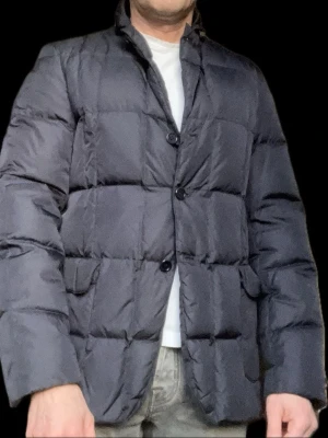 Mörkblå dunjacka från Aspesi - Mörkblå lätt dunjacka i kavajstil från Aspesi med quiltad design och knäppning framtill. Jackan har två framfickor med lock och en klassisk krage. Perfekt vårjacka. Modellen är 180 och väger 70 kg. 