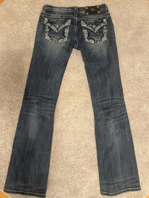 Mörkblå lågmidjade bootcut jeans från Miss Me - Snygga mörkblå lågmidjade bootcut jeans från Miss Me i storlek 26. Jeansen har dekorativa bakfickor med broderier och nitar samt klassisk femficksdesign. Midjemått: ca 37.5 cm / Ytterben: ca 103 cm / Innerben: ca 83 cm. Priset kan diskuteras! Skriv gärna vid frågor!💗