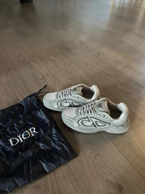 Dior b30 mörkgrå - Säljer ett par beige Dior sneakers med coola detaljer i mesh och läder. Skorna har vita och grå inslag, chunky sula och tydlig Dior-logga på sidan. Perfekta för dig som gillar exklusiv streetwear och snygg design.