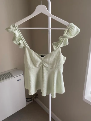 Ljusgrön volang topp med axelband - Supersöt ljusgrön topp med volangdetaljer på axelbanden och lätt v-ringad hals. Toppen har en flowy passform och är tillverkad i ett ribbat material som ger en snygg struktur. Perfekt för varma dagar och ger en fräsch look.