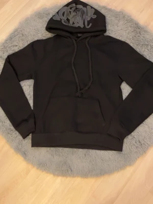 Svart hoodie med tryck på huvan - Snygg svart hoodie med stor ficka framtill och grova dragsnören. Huvan har ett grått graffiti-inspirerat tryck som ger en cool streetwear-vibe. Tillverkad i mjukt bomullsmaterial, perfekt för en avslappnad stil.