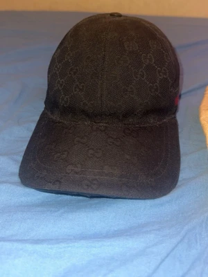 Svart keps från Gucci med monogram ÄKTA - Säljer en svart keps från Gucci med det klassiska GG-monogrammet över hela kepsen. Kepsen har böjd skärm och diskret röd-grön detalj på sidan. Tillverkad i tyg och levereras med original dustbag från Gucci.