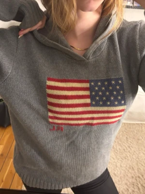 Stickad hoodie från Ralph Lauren - Säljer en grå stickad hoodie med stor amerikansk flagga på bröstet. Tröjan har huva och ribbade muddar vid ärmslut och nederkant. Tröjan är från Ralph Lauren och är vintage.
