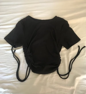 Svart tajt t-shirt med snöre  - Säljer denna svarta tajta t-shirten med snöre på sidan. Använt väldigt få gånger för många år sedan. Storlek XS och är i väldigt bra skick!🥰