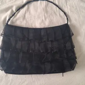 Medelstor svart handväska med fransar och volanger.Och ett litet fack med blixtlås.Vet ej ursprungspris. Väska är från Vezzano. Den är använd (Ser inte sprillans ny ut).