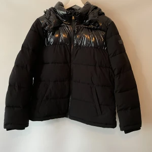 Calvin Klein jacka  - En svart puffer jacket från Calvin Klein med luva i ”shine” material.  Knappt använd, som ny.  Tre respektive 2 innerfickor (höger/vänster sida) - se bild.  Storlek Ca S. Passar mig som är 164cm.  Köparen står för frakten.