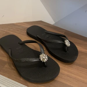 Svarta flipflops med glamig detalj - Svarta enkla flipflops med gmalig blomdetalj. Bild 3 ser du att en liten ”diamant” åkt av