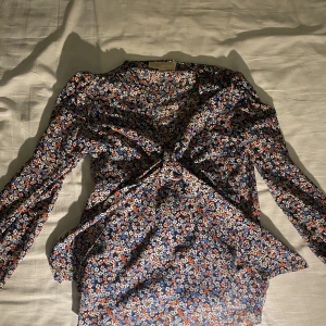 Blommig blus Michael Kors - Blus med mönster av små blommor i färgerna: blå, vitt, rött. Vid kragen finns ett band som antingen kan hänga fritt eller knytas på begäran. Ett linne är insytt och nedkommer därför. 