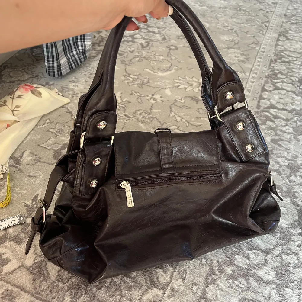Rymlig brun-svart väska från märket ”O CI GO”. Den har en ficka på baksidan, och två på insidan. Platt undersida. Använd men i gott skick! H18xB38. Accessoarer.
