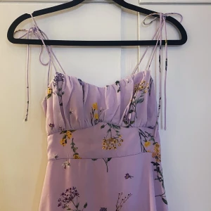 Klänning  - Vacker blommig klänning i syrenlila. Dragkedja i ryggen och axelband som knyts ihop.  Nyskick. Enbart använd två gånger 