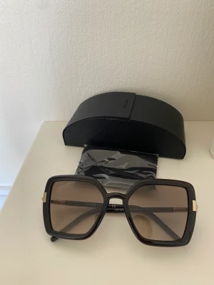 Prada solglasögon - Oanvända! Inköpta på Arlanda taxfree. Äkta. Se modellbild på andra bilden. Svart & bruna.  Fodral och torkduk medföljer. Torkduk fortfarande kvar i skyddsplast.  Gummiakiga ramar vilket gör dem bekväma.   Nypris 2200kr 