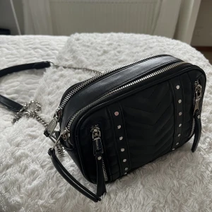 Väska - Svart väska med silvriga detaljer på. Finns två förvaringsfickor. Långt band. 