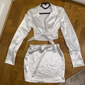 Vitt set | Belita - Ett jättesnyggt vitt set med kjol och knytbar topp. Materialet är väldigt glansigt och mjukt. Köpt i USA. Skitsnyggt att ha på sig ute på en utekväll. Säljer då kjolen är för liten för mig. Skriv för mer info och bilder.🤍 Lite nopprigt på vissa ställen!
