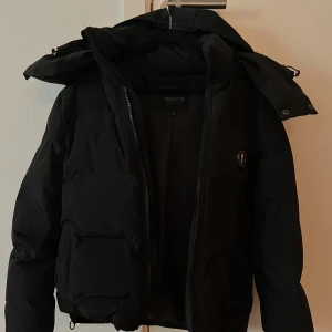 Trapstar Jacka  - Trapstar dun/vinter jacka.  Säljer för kommer ej till användning.   - Svart  - Varm  - Storlek XS (passar som S/liten M)  - Trapstar text på ryggen  - Trapstar logga på bröstet  - Resårband i midja  - Resårband i luva  - Avtagbar luva   (ÄKTA) 