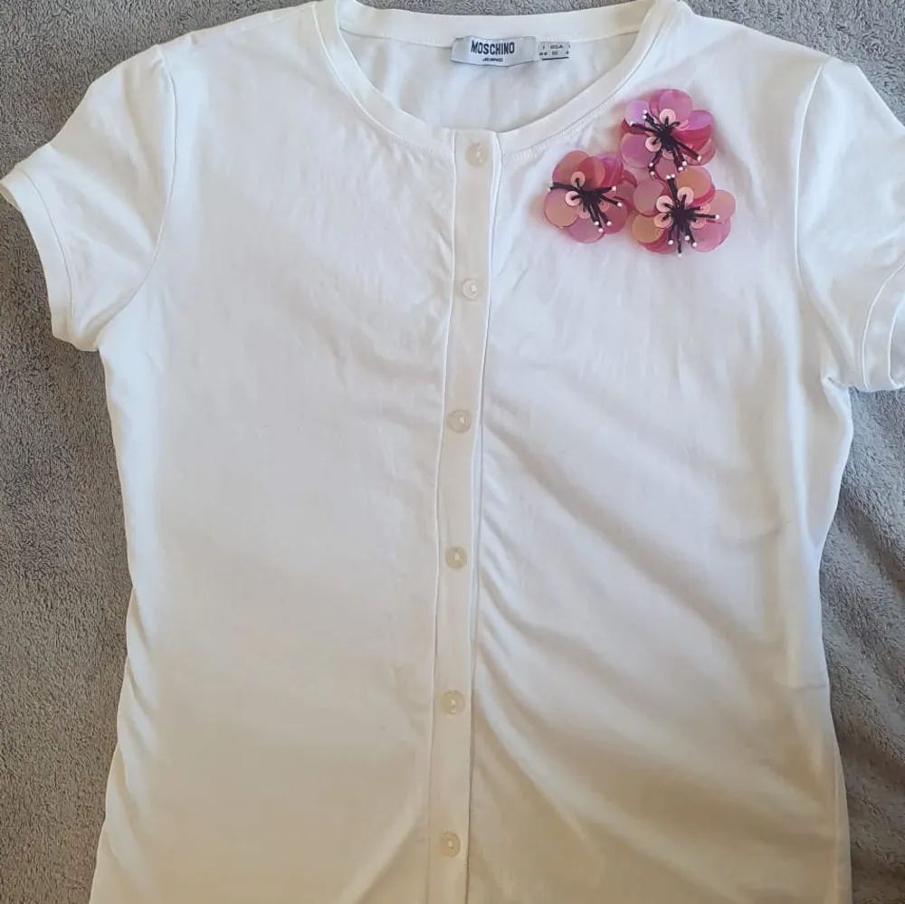 Super snygg vintage y2k T-shirt från Moschino. Super fin med rosa blomm detalj i paljett. Knappar. Italiensk 44, passar mig som har xs-s💗. Toppar.