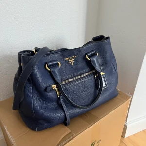 Prada väska - Väska från Prada i mörk blå med guldiga detaljer. Ett långt band ingår. Den är stor, t.ex dator får plats.