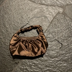 Brun liten väska - Liten väska, brun i siden-material. Både kortare handtag och längre kedja med stenar på. Nästan aldrig använd.