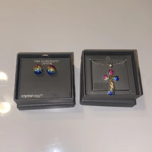 Set - Halsband och örhängen  - Silverpläterat korshalsband och örhängen med kristallstenar i olika färger.   Dessa smycken har aldrig använts och är köpta i USA.   Smyckena kommer i originalförpackning. 