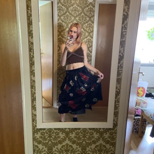 Svart blommig kjol - Svart lång kjol med röda rosor och beacha blommor. Kjolen är uppsydd 5 cm men ifall man vill ha den längre så kan man lätt sprätta upp. Storleken är 40 men jag som normalt är 36-38 kan ha den vid höfterna. 