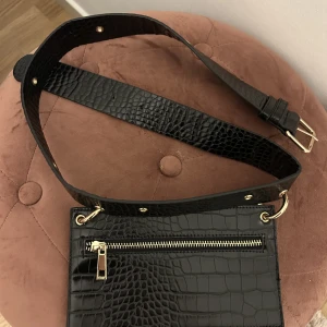 Bälte med väska  - Svart bälte med en liten väska. Imitation av ormskinn och guldiga detaljer. Går inte att ta av väskan. 