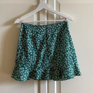Grön kjol med vita blommor - Somrig ”fake-omlott-kjol” i en fin grön färg.🍀Det finns en dragkedja på baksidan som gör det smidigt att ta på-och av sig kjolen. Materialet är skönt och den sitter väldigt fint. Hör av dig vid frågor eller funderingar. Ta hand om dig.💌