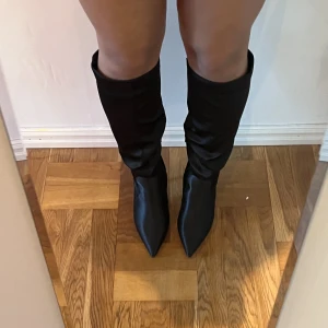 Svarta knähöga boots - Svarta knähöga boots i satin. Användna en gång.  Köpta för ca 600kr. Klackhöjd: 9,5cm Bred passform.