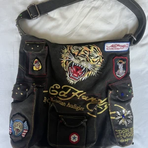 Ed hardy väska - Säljer denna Edhardy väskan med justerbart band o massa coola detaljer. Skulle tro att man får plats med dator mm i den. Fint skick, lite tecken på användning på bild 2 💓