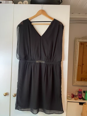 Klänning - Svart klänning med smycke i midjan. Storlek: 42. 