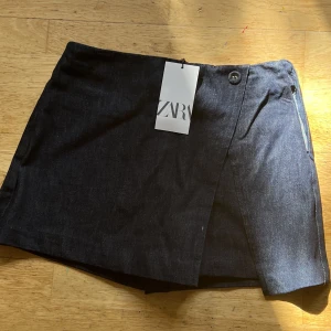 Byxkjol i denim med knapp - Jeanskjol med shorts, helt ny och oanvänd! Omlottdetalj fram, dold dragkedja Bekväm både till sommaren och hösten