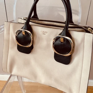 Guess väska - Beige/benvit/svart handväska från Guess. Inköpt för flera år sedan men endast använd max 3 ggr. Saknar den långa remmen, men går att haka i ny rem.  Strl bredd 34 cm, höjd 24 cm