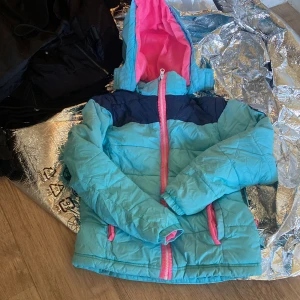 Turkos och marinblå vinterjacka med rosa detaljer - Säljer en turkos vinterjacka med marinblå axelparti och rosa dragkedja samt foder i huvan. Jackan är quiltad och har långa ärmar samt två fickor med rosa dragkedjor. Perfekt för kalla dagar!