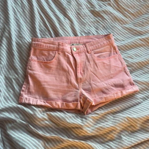 Rosa jeansshorts från H&M - Säljer ett par ljusrosa jeansshorts från H&M med klassisk femficksdesign och uppvikta benslut. Shortsen har normal passform och stängs med knapp och dragkedja framtill. Perfekta för varma dagar!