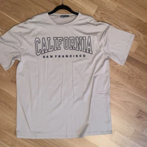 Vit t-shirt med tryck 'California San Francisco' - Vit t-shirt med svart texttryck 'California San Francisco' på bröstet. Klassisk rund halsringning och normal passform. Perfekt för en avslappnad och cool stil.