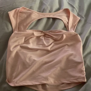 Ljusrosa croppad linnetopp med öppen rygg - Säljer en ljusrosa croppad linnetopp med breda axelband och snygg öppen rygg. Toppen har en liten rynkdetalj framtill och är tillverkad i ett mjukt, stretchigt material. Perfekt för varma dagar eller som ett lager under en skjorta.