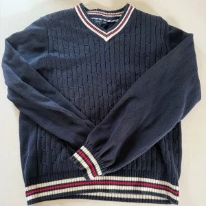 Mörkblå kabelstickad tröja med v-ringning - Mörkblå kabelstickad tröja från murrey med v-ringad hals. Tröjan har vita och röda ränder vid hals, ärmslut och nederkant. Klassisk och stilren design med lång ärm och ribbade detaljer.