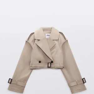 Beige croppad trenchcoat från Zara - Trendig beige croppad trenchcoat från Zara med bred krage, dubbelknäppning och bältesdetaljer vid ärmsluten. Jackan har långa ärmar och en klassisk trenchcoat-look i kortare modell.
