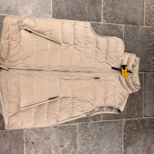 Beige quiltad dunväst från Parajumpers - Snygg beige quiltad dunväst från Parajumpers med dragkedja och två snedställda fickor framtill. Västen har en hög krage och klassisk Parajumpers-detalj vid halsen. Inga skador på västen. Mvh
