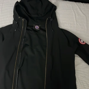 Svart vindjacka från Canada Goose med huva - Säljer en svart vindjacka från Canada Goose i storlek S. Jackan har huva, dragkedja och klassisk Canada Goose-logga på ärmen. Perfekt för blåsiga dagar och enkel att matcha med olika outfits.   2500 sek price can be discussed.