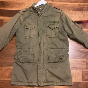 Olivgrön militärinspirerad jacka från Alpha Industries - Säljer en olivgrön militärjacka från Alpha Industries med fyra stora fickor framtill, axelklaffar och ståkrage. Jackan har dragkedja och knappar samt detaljer på ärmen. Perfekt för dig som gillar utility-stil.