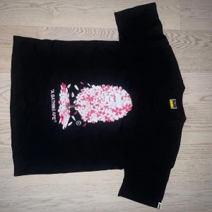 Svart t-shirt från A Bathing Ape med körsbärsblommor - Säljer en svart t-shirt från A Bathing Ape med ikoniskt apansikte täckt av rosa och vita körsbärsblommor på bröstet. Klassisk passform och rund hals. Perfekt för dig som gillar streetwear och unika prints.