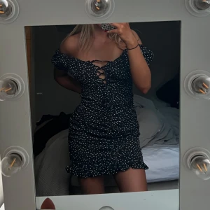 Svart kortklänning med vita prickar och snörning - Säljer en svart kortklänning med vita prickar. Klänningen har offshoulder-modell, snörning framtill och volangkant nertill. 